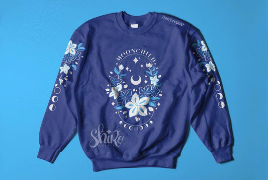 Blue sweater with Moonchild theme. Frasurbane-style central drawing with flowers and cosmic elements, Moonchild&quot text above, lunar phases below.